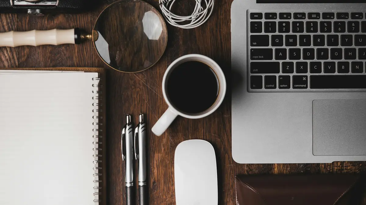 A desk seen from above with a laptop, notebook, pen and a cup of coffee